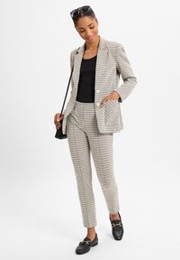 Kariertes Blazer und Hose aus beigem und schwarzem Stoff mit einer taillierten Passform. Kombiniert mit einem schwarzen Oberteil und Loafers mit Hardware-Details.