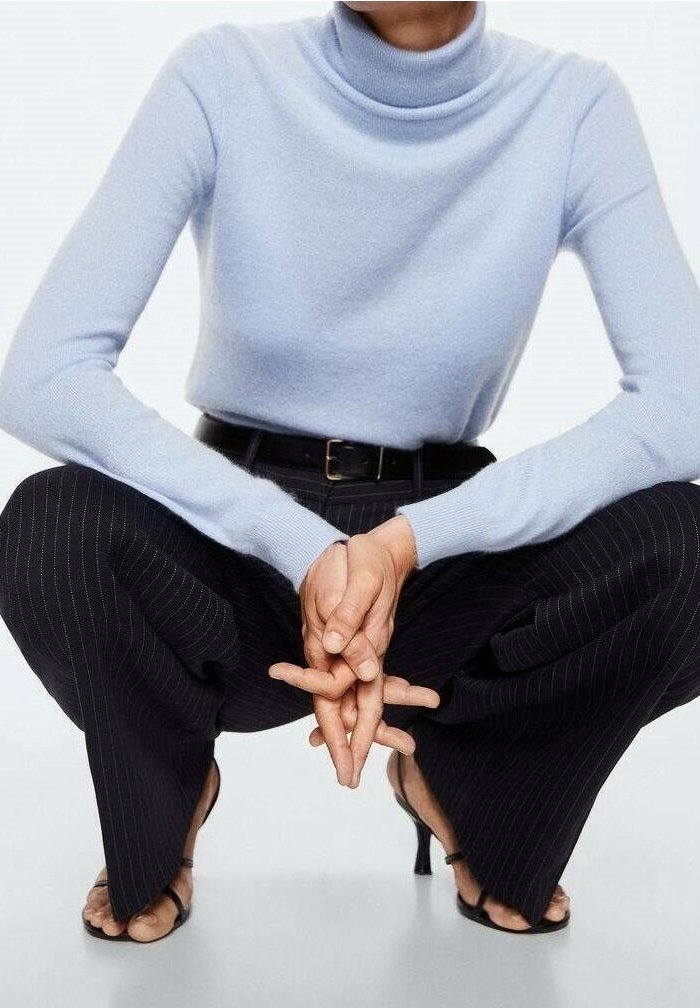 Maglione a collo alto azzurro chiaro, aderente, con maniche lunghe. Abbinato a pantaloni a gamba larga neri a righe sottili. Modella accovacciata in sandali aperti.