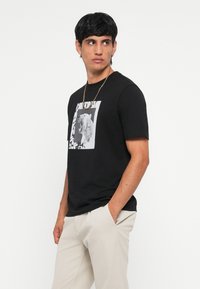 T-shirt en coton noir avec un graphique d'un lion rugissant dans un cadre blanc, associé à un pantalon de couleur claire. Tissu texturé et design à col rond.