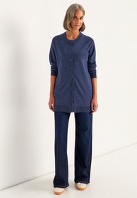 Frau mit grauen Haaren, die einen blauen, geknöpften Cardigan, eine dunkle, weit geschnittene Jeans und weiße Sneakers trägt, steht vor einem schlichten hellen Hintergrund.