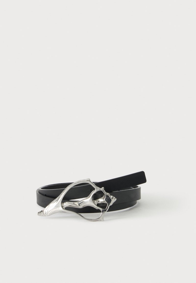 Ceinture en cuir noir avec une boucle métallique argentée abstraite en forme d'une forme organique ouverte, présentée enroulée sur une surface blanche.