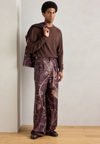 Camisa marrón de manga larga combinada con pantalones morados de gran tamaño que presentan un patrón de mapa detallado en tonos dorados. Los accesorios incluyen un bolso a juego.