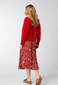 Czerwony sweter z prążkami, wzorzysta spódnica midi w czerwono-beżowe wzory o luźnym kształcie i brązowe buty wsuwane z teksturowaną podeszwą.