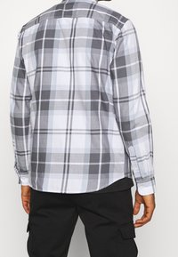 Camicia a maniche lunghe grigia e bianca a quadri con polsini a bottone, caratterizzata da diverse dimensioni dei quadri e un orlo arrotondato, realizzata in tessuto leggero.