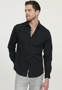 Homme à cheveux courts et barbe, portant une chemise noire à boutons avec des contours blancs et un jean gris clair, regardant vers sa gauche.