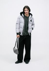 Jachetă puf gri cu glugă căptușită cu blană, accesorii negre și detaliu cu logo. Asortată cu o cămașă verde închis, pantaloni negri largi și o geantă gri de umăr.