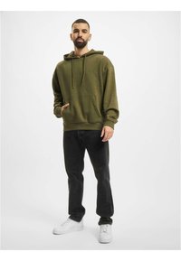 Olivengrønn hettegenser med lommer foran, teksturert materiale og snorer. Kombinert med svarte jeans og hvite sneakers.