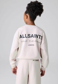 Osoba z kręconymi włosami, nosząca bladoróżowy sweatshirt i spodnie marki AllSaints, prezentująca plecy z wyraźnym czarnym napisem i japońskimi znakami.