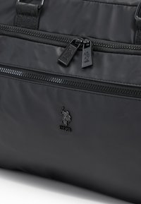 Černá nylonová sportovní taška s dvojitými kapsami na zip, tkaným popruhem a lesklým logem. Hladká textura s pevnými kovovými detaily.