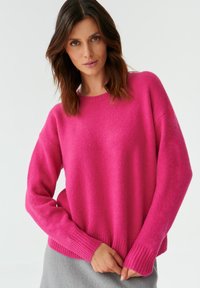 Fuchsia-Pink Pullover mit lockerer Passform, gerippten Bündchen und Saum, aus weichem Stoff gefertigt. Das Model trägt ihn über einem grauen Rock.