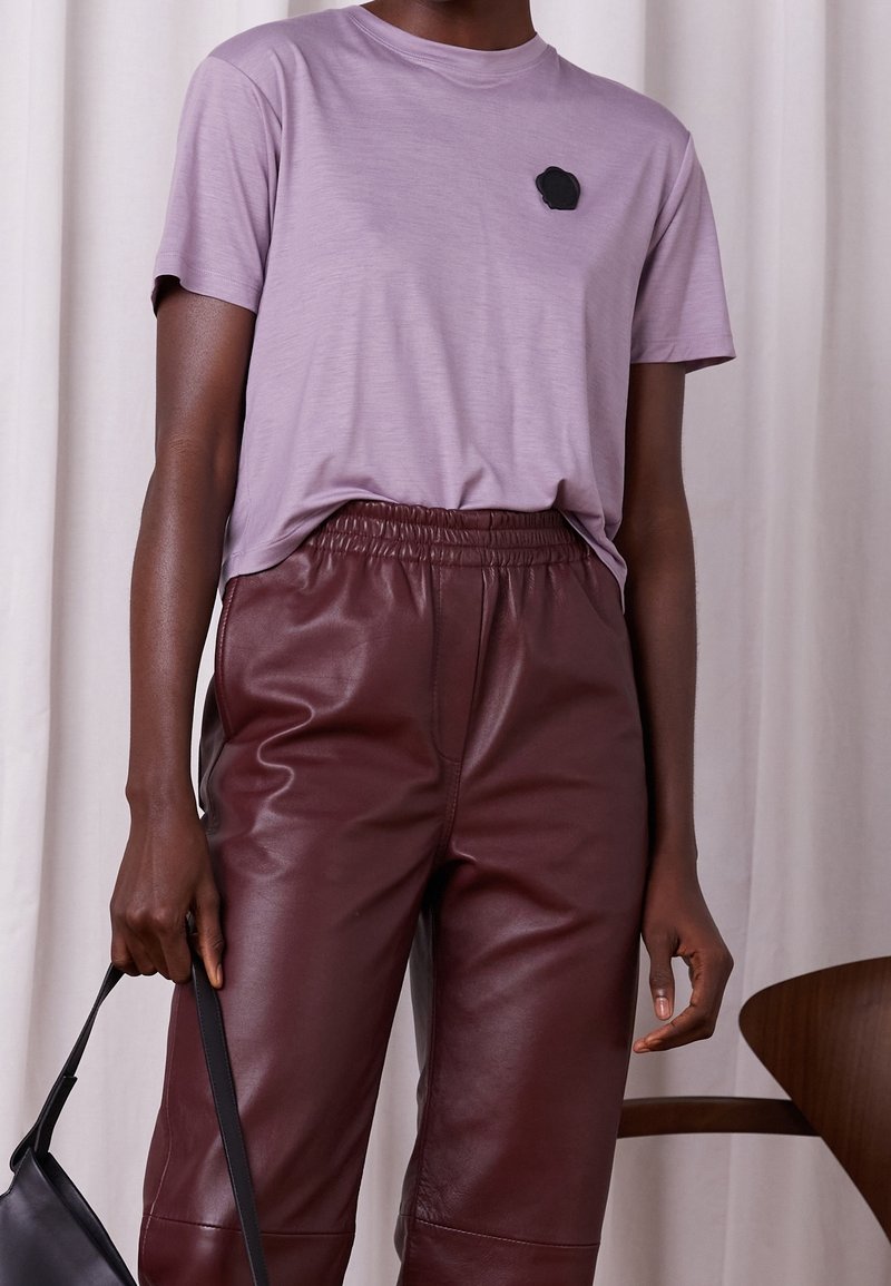 T-shirt lavande à manches courtes avec col rond et écusson noir sur la poitrine, associée à un pantalon en cuir marron à taille élastique, tenant un sac noir.