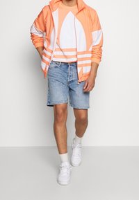 Chaqueta naranja ligera con rayas blancas, combinada con una camiseta a rayas naranja y blanca, pantalones cortos de mezclilla y zapatillas blancas.