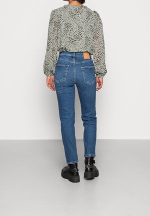 Žena oblečená v modrých džínech s vysokým pasem, světle zelené blůze s černými skvrnami a černých botách na platformě, stojící u bílé zdi.