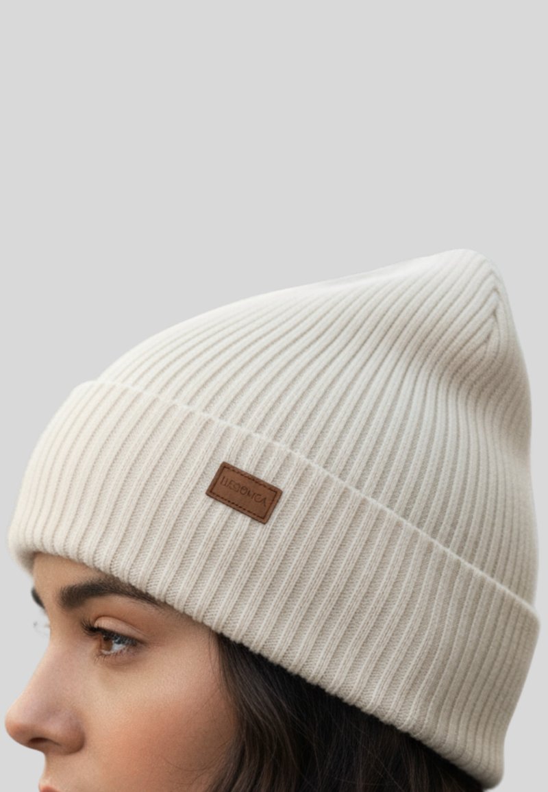 Witte geribbelde muts van gebreide stof, met een klein bruin leren label aan de zijkant en een taps toelopende bovenkant.