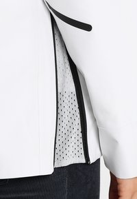 Veste blanche avec des accents noirs, présentant une doublure en mesh visible par une fermeture éclair ouverte. Tissu extérieur lisse, coupe ajustée.