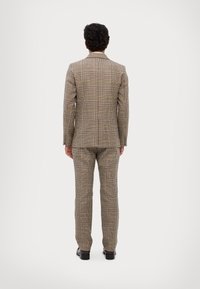 Barna kockás öltöny, amely illeszkedő sziluettel, dupla szellőzőnyílással és testre szabott nadrággal rendelkezik. Az anyag textúrált hatású; a zakó ék alakú hajtókákkal és kifinomult dizájnnal bír.