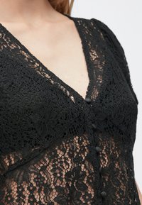Čierny čipkovaný top s výstrihom do V, zložitými kvetinovými vzormi a čiernymi gombíkmi vpredu, s priehľadným a textúrovaným vzhľadom.
