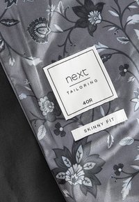 Pelēks audums ar ziedu rakstu, ar baltu birku, uz kuras rakstīts "next Tailoring 40R Skinny Fit." Ievērojama gluda tekstūra un smalkas detaļas.