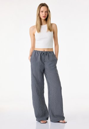 Młoda kobieta stoi, ubrana w biały, bezrękawnik krótki top, luźne spodnie w pionowe granatowo-białe paski oraz sandały z odkrytymi palcami, ręce w kieszeniach.