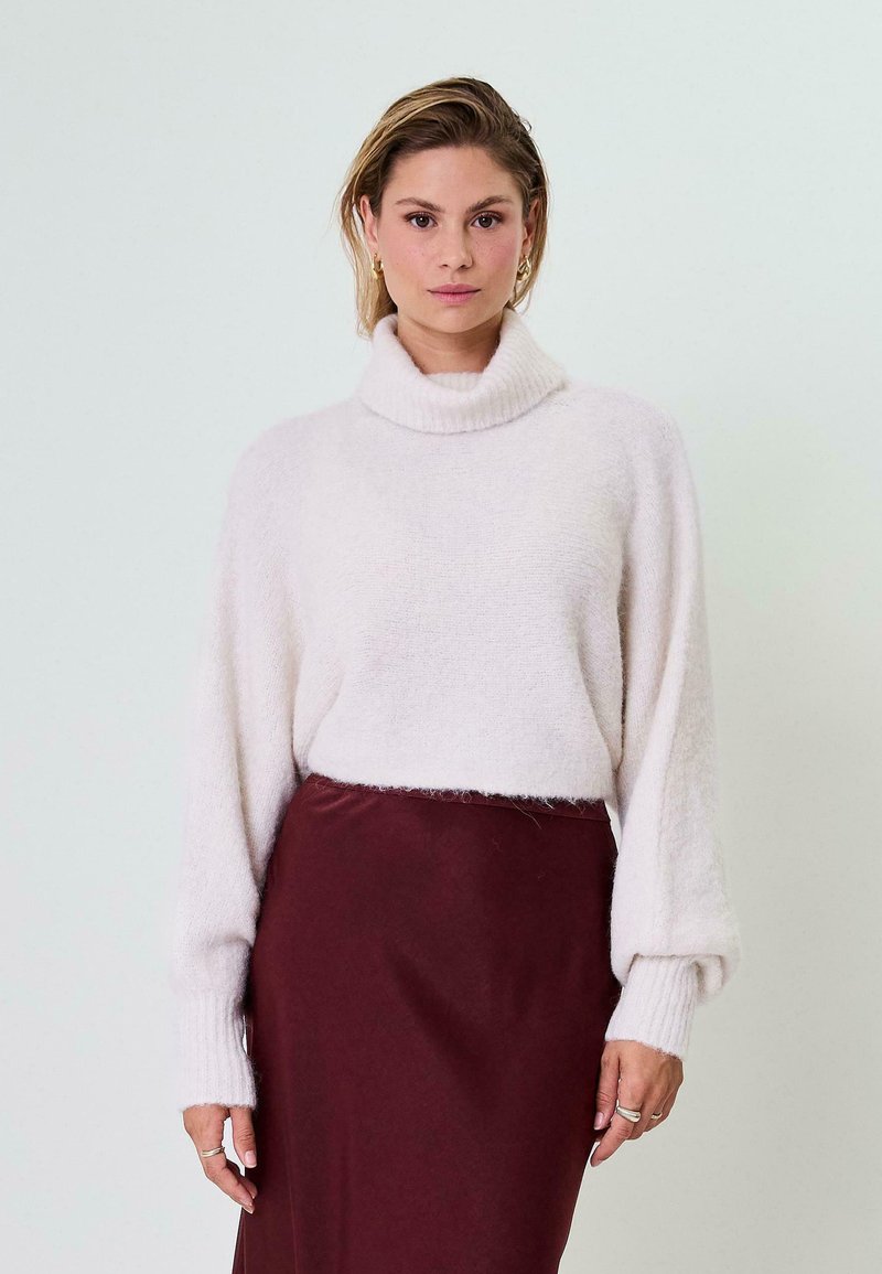 Šviesiai smėlinis turtleneck megztinis su plačiomis rankovėmis, pagamintas iš švelnios, faktūrinės medžiagos, derinamas su burgundiškos spalvos satino sijonu.