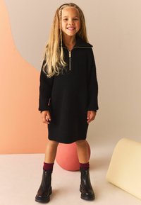 Vestito nero lavorato a maglia con collo con zip, maniche lunghe, abbinato a stivali neri con cerniera alla caviglia e calze bordeaux. Design semplice e casual.