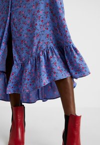 Květnaté midi šaty v světle modré barvě s drobnými růžovými a černými vzory, s volánkovým lemem a vysokým rozparkem. Vytvořeno s červenými kotníkovými botami.