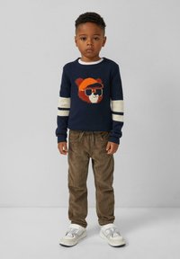 Granatowy sweter z graficznym wzorem zwierzęcia i białymi paskami. Teksturowane brązowe spodnie z korduroyu. Białe sneakersy z szarymi akcentami.