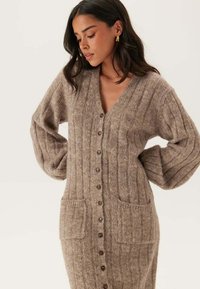 Lange beige cardigan gemaakt van ribgebreide stof, met een V-hals, knoopsluiting en twee voorkeurszakken. Zachte textuur met een ontspannen pasvorm.