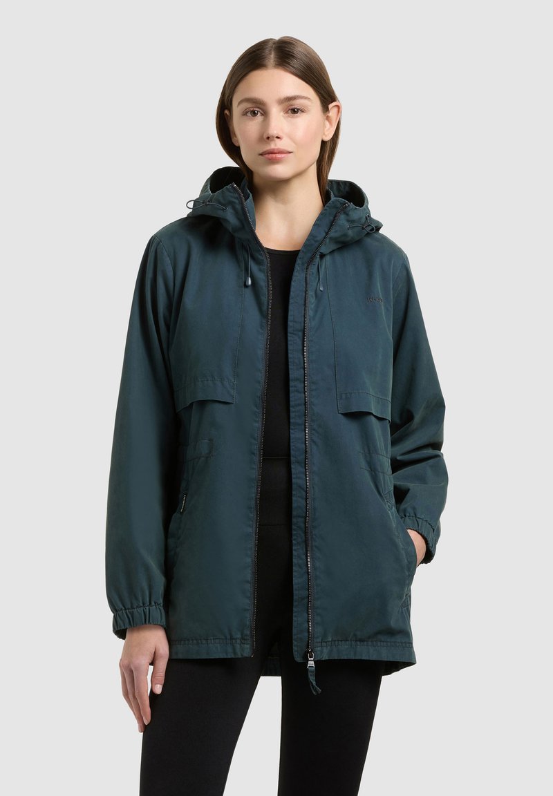 Femme portant une veste à capuche vert foncé par-dessus des vêtements noirs, debout avec une main dans la poche sur un fond gris uni.