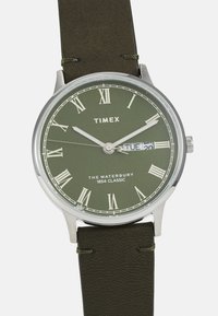 Montre à boîtier argenté avec un cadran vert, des index en chiffres romains, une fonction date et un bracelet en cuir vert olive. Bracelet texturé avec des détails de couture.