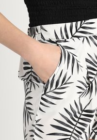 Kalhoty s lístkovým vzorem v bílé a černé barvě, vyrobené z lehkého materiálu. Obsahují boční kapsu a elastický pás pro pohodlí.