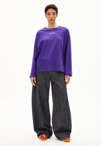Lila langärmliges Shirt mit weißem Logo auf der Brust, kombiniert mit schwarzen weit geschnittenen Jeans und orangen Sneakers.