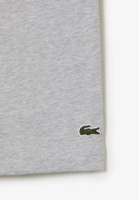 Tissu gris clair avec un petit logo de crocodile vert brodé près du bord inférieur, sur un fond blanc.