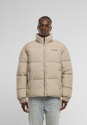 Mann iført beige vattert jakke med høy krage og glidelås, stående foran ensfarget grå bakgrunn med hendene avslappet ved sidene.