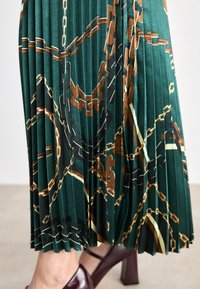 Grüne, plissierte Rock mit einem Kettenmuster in Gold- und Brauntönen. Der Stoff hat eine glatte Textur, und der Saum endet knapp über dem Knöchel.