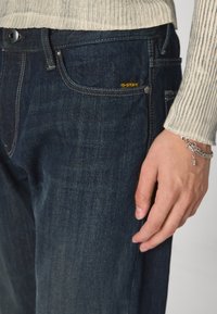 Temno modri kavbojke z ožjim krojem, ki imajo majhen rumen G-Star logotip na sprednjem žepu in kontrastne šive. Vidni so rebrasti manžeti puloverja.