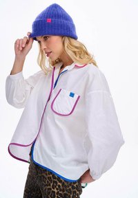 Biała koszula z guzikami, z różowymi i niebieskimi lamówkami, przednią kieszonką i objętołowymi rękawami, w połączeniu z fioletową dzianinową czapką.