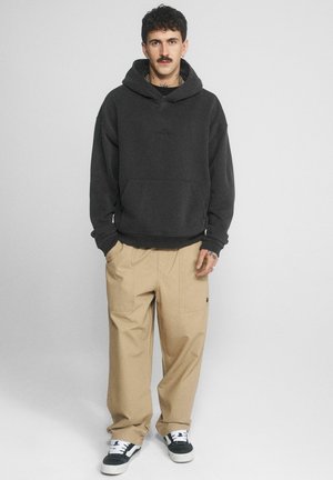 Mężczyzna z wąsami w czarnej bluzie z kapturem, beżowych szerokich spodniach i czarno-białych trampkach stoi na gładkim jasnym tle.