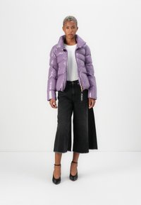 Lilla pufferjakke med en blank finish, høj krave og guldfarvet hardware, parret med sorte bredbenede jeans og sorte hæle.