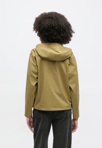 Αδιάβροχο τζάκετ ελιάς με κουκούλα, με λεία υφή και άνετη εφαρμογή· φορεμένο με παντελόνι σκούρου γκρι, με ελάχιστες λεπτομέρειες.