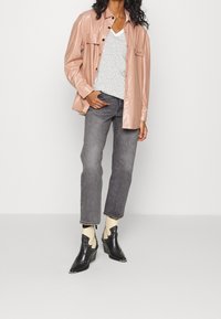 Camisa chaqueta de cuero sintético de color rosa claro, blusa blanca de escote en V con puntos, jeans rectos grises y botas negras de tobillo con recortes y tachuelas.