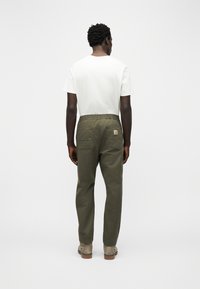 Άνδρας που στέκεται με την πλάτη προς το μέρος μας, φοράει λευκό μπλουζάκι, χακί παντελόνι με πίσω τσέπες και λογότυπο, και μπεζ μπότες.