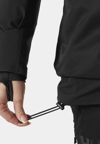 Czarna kurtka odporna na warunki atmosferyczne, na której widać rękę dostosowującą sznurek ściągający na dole. Materiał ma gładką, matową teksturę.