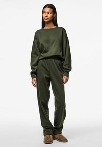 Olivgrüner Sweatshirt und passende Jogginghose aus weichem Stoff, mit lockerer Passform und gerippten Bündchen, kombiniert mit hellbraunen Hausschuhen.