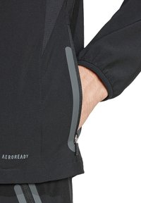 Musta kevyt takki harmailla yksityiskohdilla ja vetoketjullisilla taskuilla. Ominaisuuksiin kuuluu teksturoitu kangas ja istuvat hihansuut. Brändin nimi "AEROREADY" näkyvissä.