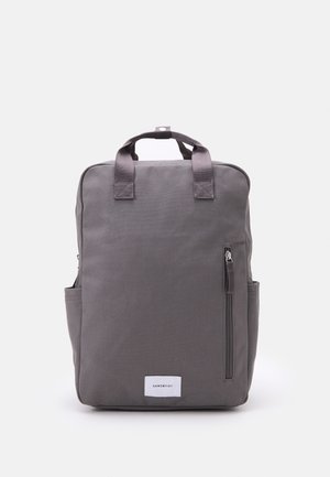 Grå tygryggsäck med dubbla topphandtag, en framficka med dragkedja, sidofickor och en logotyp-patch längst ned. Slät textur och rektangulär form.