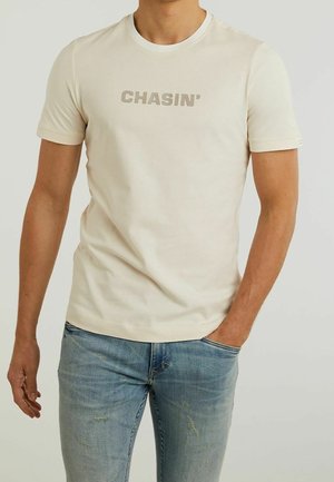 T-shirt beige clair avec le texte "CHASIN'" en gris subtil. Col rond et manches courtes. Porté avec un jean bleu clair texturé.