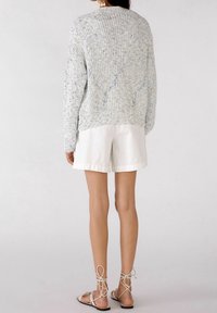 Person som bär en ljusblå prickspräcklig stickad tröja, vita shorts, beiga snörsandaler, som står mot en neutral vit bakgrund, med ryggen vänd. 