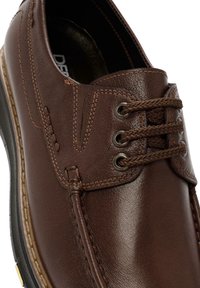 Chaussure en cuir marron avec un bout arrondi, surface texturée, couture décorative et œillets en métal pour les lacets. Semelle intérieure rembourrée visible.