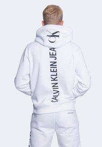 Sudadera blanca con capucha que presenta un gran logo negro de "CALVIN KLEIN JEANS" en vertical a lo largo de la espalda. Tejido suave y ajuste relajado.
